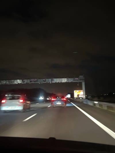 Night scene on a highway with cars driving in both directions. A bridge runs parallel with the highway, partially obscured by darkness. Advertisements are visible above the bridge. Car headlights and taillights illuminate the road. The image is taken from inside a vehicle, showing part of the vehicle's interior on the right side.