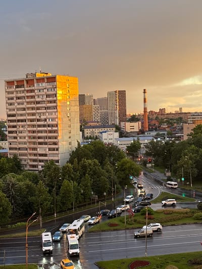 The image captures an aerial view of a city intersection in Mytishchi, Russia, during a dusky sunset. The sky is a mix of grey and warm orange hues, suggesting either pre-dusk or post-rain conditions. The foreground is dominated by a wet, multi-lane road reflecting the ambient light, with several vehicles present.

On the road, a yellow taxi is stopped at a pedestrian crossing. Further ahead, a white bus and two white vans are navigating the intersection. To the left, a dark-colored car is turning, and behind it, other cars are visible. A white SUV is positioned in the right lane, and a light-colored sedan is turning right. A circular island with green bushes and red flowers is situated within the intersection.

The middle ground features a dense canopy of green trees lining the roads and surrounding the buildings. Several buildings are visible, including a tall, light-colored apartment building with balconies on the left, illuminated by the setting sun. In the background, a reddish-orange and white striped industrial chimney stands tall. Other buildings, including modern high-rises and lower industrial structures, are scattered across the cityscape. Traffic lights are visible at the intersection, some showing green signals. Streetlights are also present along the roads. The overall scene depicts a typical urban environment experiencing the transition from day to evening.