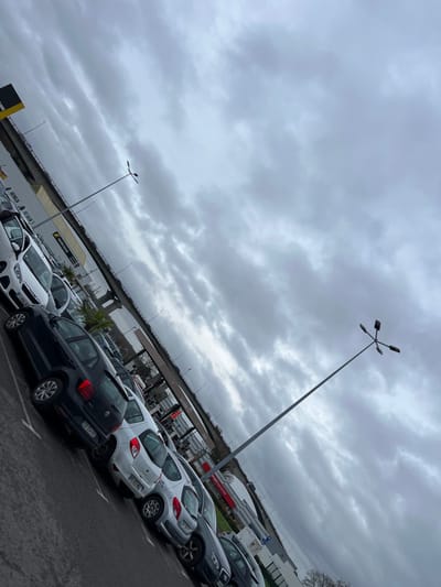This image depicts an outdoor parking lot under an overcast sky, taken with a pronounced tilt from the bottom-left to the top-right. The location is identified as Hérouville-Saint-Clair, France, specifically in a commercial area near an elevated road.
In the foreground and midground, numerous passenger vehicles are parked in rows on an asphalt surface. The vehicles include various hatchbacks and sedans, predominantly in white, black, and silver/grey colors. A white Peugeot 208 is visible on the far left, followed by a black Volkswagen Polo, and several other compact cars. Some vehicles have French license plates visible (e.g., ES-554-CL on the black Polo, ES-854-HX on a white hatchback). The asphalt appears damp in areas.
Two tall, grey lampposts with multiple light fixtures (four per pole, arranged radially) extend upwards. The one in the far right midground is particularly prominent, reaching towards the upper right corner of the frame.
In the background, an elevated road or bridge, supported by grey concrete pillars, traverses horizontally across the scene. Below the elevated road and behind the parked cars, several commercial buildings are visible. A yellow sign for "NORISKO Auto" (a French vehicle inspection network) is prominent on the left. Further to the right, a fuel station canopy with red signage and several pump islands is identifiable. A digital display at one of the pumps shows red numbers, possibly indicating fuel prices (e.g., "1.898"). Beyond the fuel station, a building with a red facade and a white sign partially visible as "LE PREM" is seen. Sparse vegetation, including a palm-like plant and bare trees, is present around the commercial structures.
The sky is entirely covered with dense, grey clouds, indicating an overcast day. No individuals are visible in the scene, and no specific actions or interactions are depicted. The overall impression is one of a typical commercial parking area on a dull, possibly damp, day.