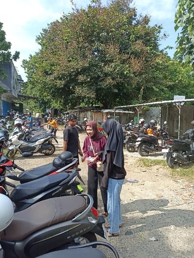 This media file captures an outdoor scene, likely a motorcycle parking area in Mataram, Indonesia, during bright daylight.
**Subjects:** The image features numerous people and many motorcycles. In the foreground, three individuals are central: two young women and a young man. The woman on the left wears a maroon long-sleeved shirt and a light brown hijab, while holding a small item. The woman on the right wears a black top, light blue jeans, and a black hijab. The man stands between them in a black t-shirt and dark shorts. Several other people are visible in the background, mostly near motorcycles.
**Objects:** The most prominent objects are the many motorcycles and scooters of various models, parked closely together, filling a large portion of the frame. Helmets are seen on some motorcycle seats. A large, dense-foliage tree dominates the upper background, along with other greenery. Buildings with some blue details are visible on the left side, and low, possibly makeshift structures or fences line the background. The ground is unpaved dirt and gravel.
**Activity or event taking place:** The central group appears to be engaged in a casual conversation, possibly sharing or discussing something, as one woman holds an item. The broader scene depicts a busy motorcycle parking lot, suggesting people are either arriving, departing, or waiting.
**Notable details:** The time of day is clearly mid-day or early afternoon, evidenced by the bright sunlight and distinct, strong shadows cast by people and objects. The weather is sunny and clear. The location appears to be an informal outdoor parking area, likely for a public space or educational institution given the number of scooters, consistent with the specified context of Mataram, Indonesia. The clothing, including the hijabs worn by the women, aligns with local attire. Emotions seem neutral to pleasant, with the central figures engaged in conversation.
**Visible text:**
* The word "CHILL" is printed on the maroon shirt of the woman on the left.
* "HONDA" is visible on the side of a white scooter in the midground.
* "SCOOPY" is visible on the side of a dark scooter in the foreground.
* Some indistinct white text is visible on a green sign in the background.