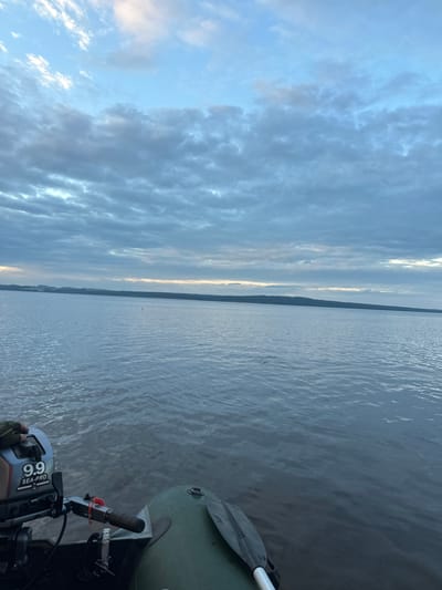 The image depicts a serene water scene, likely taken from a small motorboat. The foreground shows a portion of a green inflatable boat with a silver outboard motor that reads "9.9 SEA-PRO". A hand wearing a green glove is visible resting on the motor. A paddle lies in the boat. The water is calm with gentle ripples, reflecting the overcast sky. In the distance, a long, dark landmass, possibly a forested shoreline, stretches across the horizon. The sky is filled with a dramatic array of gray and white clouds, with a faint band of lighter sky near the horizon, suggesting either sunrise or sunset. The overall atmosphere is tranquil and natural.