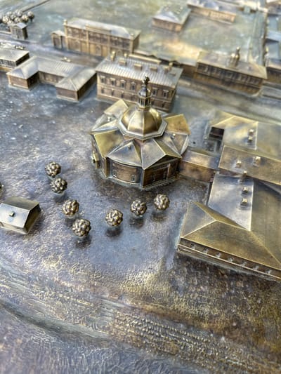 This is a close-up, high-angle, eye-level shot of a bronze architectural model. The model depicts a city with multiple buildings and a central circular structure topped with a dome and a spire. The buildings are intricately detailed, showing roofs, windows, and entrances. The ground surface of the model is textured, resembling cobblestones or uneven terrain. Scattered across the ground are several small, spherical objects that appear to be stylized trees or bushes. In the lower left quadrant of the image, a small, rectangular building with a pitched roof is visible. The lighting is natural, casting subtle shadows and highlighting the metallic sheen of the bronze. The model is located in Barnaul, Russia, and represents the area surrounding the Barnaul Ascension Cathedral.