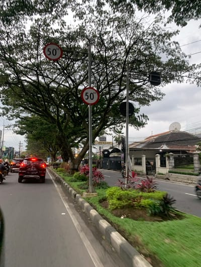 The image shows a street scene in Malang, Indonesia, located at latitude -7.94 and longitude 112.637. A black SUV is traveling on the road in the middle of the frame. There is a row of trees on the left side of the road, and a curb with green grass and bushes. Two round speed limit signs reading "50" are visible on a pole on the left side of the image. A building with a white facade is visible in the background. The road surface is black asphalt with white markings. The sky is cloudy, and the scene is captured during daytime. A motorcycle is visible in the far right corner of the image, traveling in the opposite direction of the SUV.
