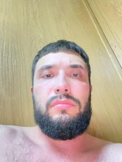 A close-up, low-angle, selfie-style photograph shows a partially cropped, fair-skinned adult male with dark hair, dark eyebrows, and a full, dark beard. The man is looking directly at the camera. His eyes are open, and his lips are slightly parted. The background consists of a light-colored, textured surface that appears to be wood paneling, with vertical lines suggesting a door or partition. The lighting in the scene is bright, casting some subtle shadows and highlighting the textures of the wood and the man's skin. The man is shirtless, with visible hair on his chest and shoulders. The image appears to have been taken indoors. There is no discernible information about the location within the city of Unknown, Bangladesh.