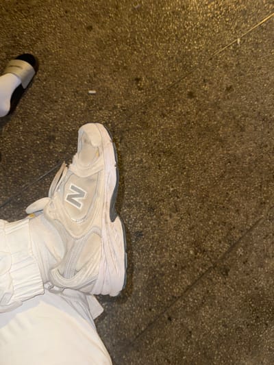 The image depicts an overhead view of ground level in Azor, Israel. In the lower-left, a left foot is visible, clad in a light-colored, worn New Balance sneaker. The sneaker displays a prominent "N" logo and shows signs of dirt and scuffing on its sole and upper. The foot is partially covered by loose-fitting, light-colored fabric, likely pants. To the upper-left, a second foot is partially visible, wearing a white sock and a black slide sandal with a gold-colored strap. The ground consists of dark, speckled concrete or asphalt, exhibiting a faint diagonal seam or crack. A single white cigarette butt rests on the ground surface, positioned near the upper-mid-left of the frame. The scene is illuminated, suggesting ambient or artificial light. No active interactions or specific actions are discernible beyond the static presence of the footwear and ground.
