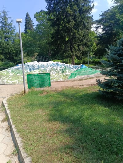 This media file shows an outdoor scene in Sandanski, Bulgaria. The main subject is a model of Pirin Mountain, a mountain range in southwestern Bulgaria, part of the Rila-Rhodope massif. The model depicts the mountainous terrain with white peaks and green lower slopes, surrounded by a decorative metal fence.
In front of the mountain model, there is a green informational sign with text in Bulgarian. The sign explains that Pirin Mountain is located in Bulgaria, covers 2585 sq km, and its highest point is Vihren peak at 2914 meters. It also highlights the mountain's rich water resources, with 186 lakes, and its diverse flora and fauna, including edelweiss, wild goats, bears, and the golden eagle. The sign mentions that Pirin National Park was established in 1962 to protect this natural beauty.
A tall lamppost with a white light fixture stands to the left of the model. The scene is set in a park-like environment with lush green grass covering the foreground and grass patches along a concrete path. In the background, tall, mature trees, including conifers and deciduous trees, create a dense forest canopy against a clear blue sky, suggesting a sunny day. The overall atmosphere is peaceful and educational, likely intended for visitors to learn about the local geography and nature. No people are clearly visible in the image.