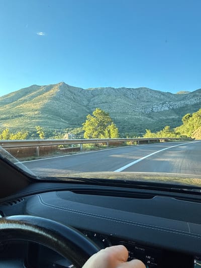The image captures a driver's perspective from within a car, traveling on a road that winds through a mountainous landscape. The road, paved with asphalt, features a white dividing line. Guardrails made of metal line the side of the road. Lush green vegetation covers the slopes of the mountains, which are bathed in the warm, golden light of what appears to be late afternoon or early morning. A clear blue sky with a faint, elongated cloud is visible above. The interior of the car, including the steering wheel and dashboard, is partially visible in the foreground. A hand is holding onto the steering wheel. A digital display on the dashboard shows information including "Від старту" (From start), "1:33 год" (1:33 hours), and "39 км/год" (39 km/h). The scene is likely in Stupovi, Montenegro, given the characteristic mountainous terrain and vegetation.