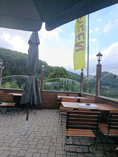 This is an outdoor seating area with tables and chairs, presumably for a restaurant or cafe, with a view of a wooded hillside. The foreground shows a paved area with several wooden tables and benches. A large, closed umbrella is prominently displayed in the center of the image, its dark fabric contrasting with the lighter pavement. In the background, a yellow flag with text is visible, fluttering on a pole. Several lampposts are also present, adding to the atmosphere. The backdrop is a lush green hillside covered in trees, suggesting a natural or semi-rural setting. The sky is partly cloudy. The overall impression is one of a peaceful, possibly quiet, outdoor space.

This scene is located in Hallein, Austria, specifically at the Gasthof zum Dürrnberg.