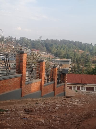 This image captures a daytime scene in Kigali, Rwanda, featuring a security fence and structures set against a hilly, vegetated landscape.

In the foreground, a reddish-brown dirt path or unpaved surface, scattered with minor debris, extends across the lower portion of the frame. A security fence, running diagonally from the lower-left to the mid-right, dominates the mid-foreground. This fence is constructed with regularly spaced reddish-orange brick pillars and a lower brick wall, accented by a gray concrete base and cap. Dark-colored metal grilles with angular patterns are installed between the brick pillars. Coiled razor wire (concertina wire) is affixed along the top of the entire fence structure.

Behind the fence, in the midground, a building with a terracotta-tiled roof and light-colored walls, featuring multiple rectangular windows, is partially visible on the right. To the left, further up the sloping terrain, several other buildings, primarily residential in appearance, are discernible amongst partial vegetation.

The background is dominated by a densely forested hillside covered with various green trees. The sky above is a pale blue with diffuse light cloud formations. No individuals, discernible actions, or specific interactions are depicted within the scene. The environment suggests an urban or peri-urban area integrated with natural topography.