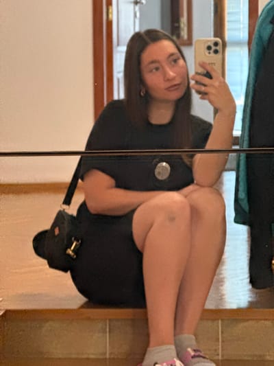 A young woman with long, dark hair is taking a selfie in a mirror. She is wearing a black t-shirt and black shorts, with grey socks. She is sitting on wooden steps inside a building. A black purse with gold hardware is slung over her shoulder. The background appears to be an interior space with wooden trim and a door. The location is not specified as Sozopol, Bulgaria.