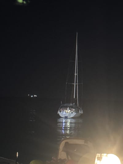 The image captures a sailboat at night on the water, likely in the area of Balchik, Bulgaria. The sailboat is illuminated, with lights visible on its deck, suggesting the presence of people. The water reflects the lights from the boat and other distant lights, creating a shimmering effect. The background is dark, indicating it is nighttime. In the foreground, there appears to be another boat or structure with some lighting, partially obscuring the view of the sailboat. The overall scene suggests a peaceful evening on the water.