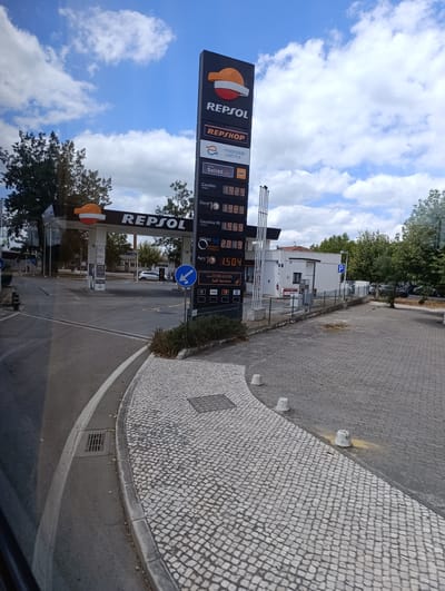 The image shows a Repsol gas station in Salvaterra de Magos, Portugal. A tall sign displays fuel prices, including Gasoleo Simples at 1.929, Diesel Color at 1.989, Gasolina 95 at 1.969, Gasolina 95 Premium Formula at 2.019, and Agro at 1.504. There is also information about "e-solred mobilidade elétrica" and "dkv". The gas station canopy, branded with the Repsol logo, is visible in the background, along with a white car. The surrounding area is paved with cobblestones and asphalt, with some greenery and trees. The sky is blue with scattered clouds. There are no people clearly visible in the image. The scene appears to be a typical urban or semi-urban setting.