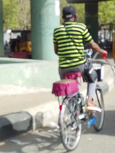 A single individual is depicted from behind, riding a bicycle on a paved road in the city of Gandu Albasa, Nigeria. The person wears a black baseball cap, a short-sleeved shirt with horizontal black and lime green stripes, grey trousers, and light brown slip-on shoes. A black shoulder bag is worn across their back. The bicycle features a dark frame, silver wheels, a prominent rear rack equipped with a dark pink/maroon padded passenger seat or cushion, and red handlebar grips. A red reflector is visible on the rear fender. The scene includes a light green concrete barrier or curb running parallel to the road on the left. In the background, large light green cylindrical pillars support an elevated structure, with blurry yellow and red vehicles and green vegetation visible beyond them. Dark grey concrete blocks are present on the ground to the left of the bicycle's path.