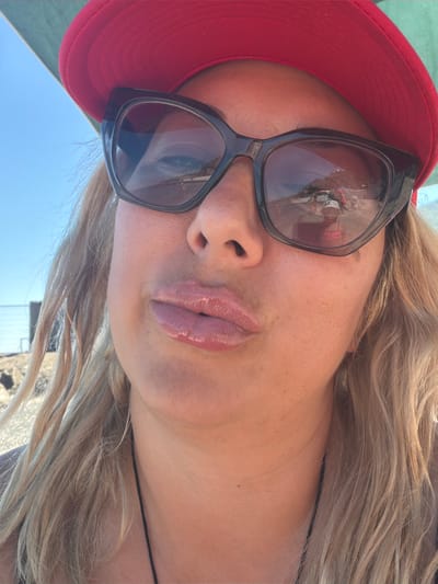 In an outdoor, daytime scene, a person with blonde hair, wearing a red hat and dark sunglasses, is seen in a close-up shot. The person's lips are slightly pursed. The sunglasses reflect a beach scene with several people and a body of water. The background shows a clear blue sky. There is no indication of the specific location within Partenit, Ukraine.