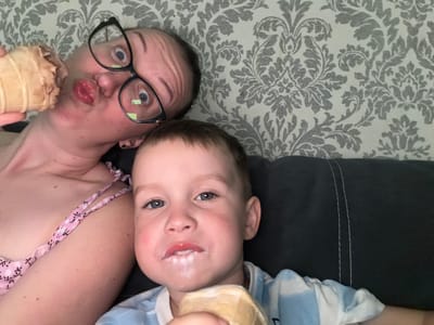 A selfie is taken from a slightly elevated angle, showing a woman and a young child enjoying ice cream. The woman is on the left, lying down and holding an ice cream cone to her mouth, her lips pursed. She wears black-rimmed glasses and a pink top with a floral pattern. Her eyes are wide, looking directly at the camera.

To her right, the child is also lying down, facing the camera with an ice cream cone in his right hand, partially covering his mouth. He has white residue from the ice cream around his lips and chin. He wears a blue and white striped shirt. The background consists of a grey and white floral patterned wallpaper. The overall impression is one of casual, shared enjoyment. The image does not provide any specific geographical indicators to confirm its location in Bila Tserkva, Ukraine.