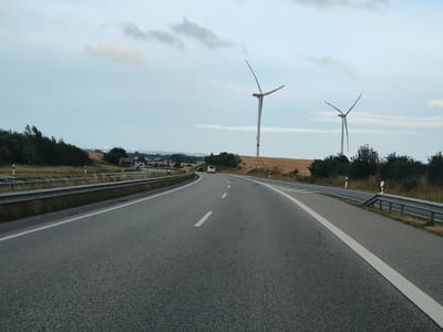 A view from a vehicle on a highway approaches a bend. Two wind turbines with red accents on their blades are visible in the distance, standing in a field. The highway has a concrete barrier on the left and a metal guardrail on the right. Several vehicles, including a white van and a car, are on the highway ahead. The sky is cloudy and overcast. In the far distance, a low-lying town or city with scattered buildings and more wind turbines is visible on the horizon, suggesting the location is near Heiligenhafen, Germany. The surrounding landscape consists of fields and trees.