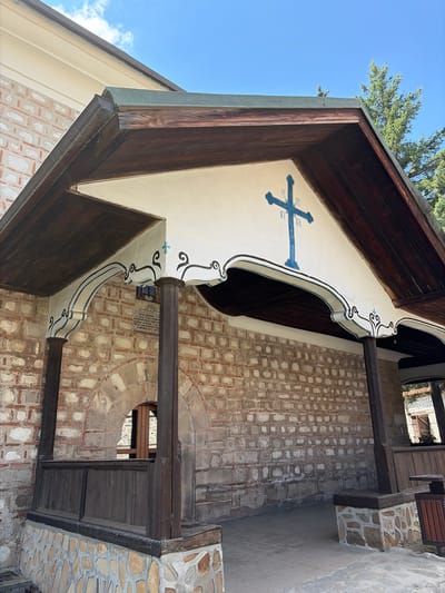 The image shows an exterior view of a building with a pitched roof supported by wooden beams. The building's facade is constructed of stone and brick, with arched openings. A blue Christian cross is painted on the white wall beneath the roof's overhang, accompanied by decorative blue and black scrollwork. A plaque with text is mounted on the wall between two wooden support posts. Wooden railings and stone foundations are visible at the lower levels of the building. In the background, a tall evergreen tree stands against a clear blue sky with scattered clouds. This is a detailed view of the entrance to the Holy Trinity Church in Bansko, Bulgaria, a significant landmark in the town.