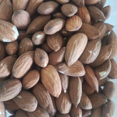 The image is a close-up, overhead shot of a pile of almonds. The almonds are a light brown color and are irregularly shaped, with some appearing more oval and others more elongated. Some of the almonds have visible texture lines, while others are smoother. A few almonds show small white imperfections or specks. The lighting is even, suggesting an indoor setting, and the background is a light, neutral color, possibly a table or countertop. There are no people, actions, or interactions depicted in the image. The image is a static, detailed view of a food item and does not provide any information about a specific location in Los Teques, Venezuela, or any other geographical area.