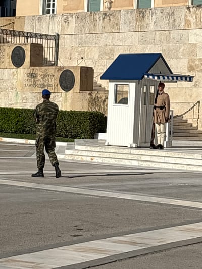The image depicts the Monument of the Unknown Soldier in Syntagma Square, Athens, Greece. Two soldiers are present. In the foreground, a soldier in a camouflage uniform and a blue beret stands with his back to the camera, facing towards the monument. In the background, a second soldier, dressed in a traditional Evzone uniform with a tan tunic, white leggings, and a red fez, stands at attention by a white sentry box with a blue roof. The monument is constructed of stone blocks, with inscriptions and circular medallions visible on its facade. A green hedge forms a backdrop behind the soldiers. The ground in front of the monument is paved with asphalt and marked with white lines. Stairs lead up to the monument platform.
