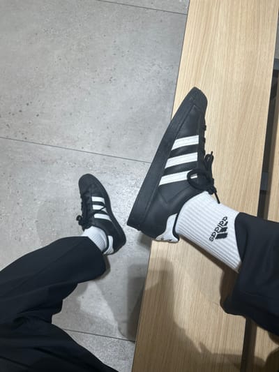 The image provides a low-angle, downward perspective of a person's lower legs and feet in an indoor environment, located in Garston, United Kingdom. The subject wears black trousers. Both feet are clad in white ribbed crew socks, with the right sock displaying a black Adidas logo. The individual wears black Adidas sneakers, each featuring three prominent white stripes on the lateral side and white accents on the heel tab. The left foot rests flat on a light grey tiled floor, characterized by visible grout lines. The right foot is elevated, with the shoe's sole resting on a light-colored wooden surface, consistent with a bench or plank. The sneakers possess black laces and solid black soles. No other individuals or specific environmental markers beyond the floor and wooden surface are visible.