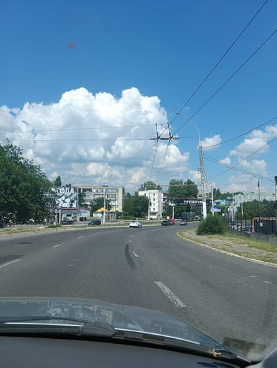 A street view from within a vehicle traveling on a road in Bender, Moldova, reveals a sunny day with a bright blue sky dotted with fluffy white clouds. The road is asphalt with white lane markings and tire tracks visible. Buildings of various sizes and architectural styles line the sides of the road, interspersed with trees and advertising billboards. Some of the billboards display text in Cyrillic. Utility poles and overhead wires, likely for public transportation, cross the sky. Several cars are visible on the road and parked along the side. The reflection of buildings and trees can be seen on the vehicle's windshield. The scene depicts a typical urban streetscape on a clear day.