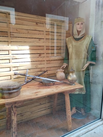 A mannequin clad in a medieval green tunic with gold trim and headdress stands behind a rustic wooden table. The table displays a collection of period artifacts: a pot, a pitcher, a metal bowl, and a two-handed sword resting on a scabbard. The setting is within a shop window, with a textured wooden wall visible in the background and a tile floor.