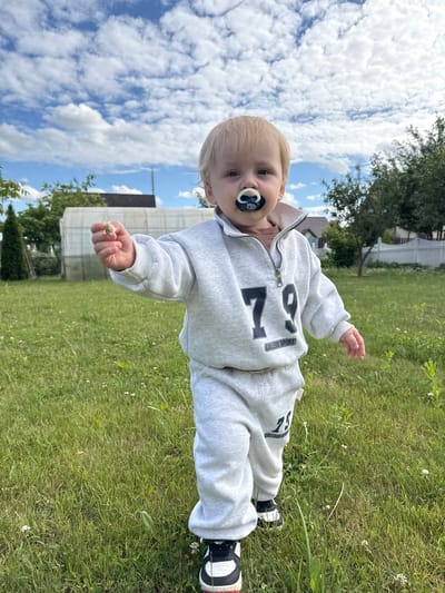 In a residential area of Gvardeysk, Russia, a toddler with blond hair stands in a grassy yard under a partly cloudy sky. The child is wearing a light gray tracksuit with the number "79" printed on the front and a pacifier in their mouth. They are holding a small white flower in their right hand and looking towards the camera. The background features a greenhouse, a white picket fence, and trees. The overall scene suggests a casual outdoor activity for a young child.