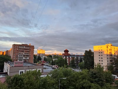 The image shows a cityscape view in Elektrostal, Russia, taken from an elevated position. In the foreground, there are green trees and rooftops of lower buildings. Beyond those, there are several multi-story residential buildings. One building on the right is reflecting the warm light of the setting sun. There is a distinctive red brick building with a tower-like structure in the middle ground. In the background, there are more buildings, some also illuminated by the setting sun. The sky is filled with gray and white clouds, suggesting either early morning or late afternoon. Two powerlines stretch across the upper portion of the image.