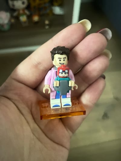 A person's hand is holding a LEGO minifigure. The minifigure is standing on an orange LEGO baseplate and appears to be a representation of a scientist or researcher, possibly holding a tablet or laptop. The figure has brown hair, a tan face, and is wearing a pink jacket over a blue shirt and striped pants. The person's fingernails are visible and appear to be painted. The background is blurred and shows what might be shelves with other figures or toys. The image does not provide any information about the city of Kommunarka, Russia.
