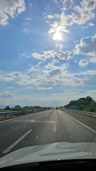 The image shows a highway with cars driving under a bright, sunlit sky with scattered clouds. The perspective is from inside a vehicle, looking forward. The road is asphalt with clear lane markings. Guardrails are visible on the sides of the road, and vegetation lines the perimeter. A green road sign is visible on the right side of the highway, indicating distances to Sofia and 112. The overall impression is a sunny day traveling on a highway.