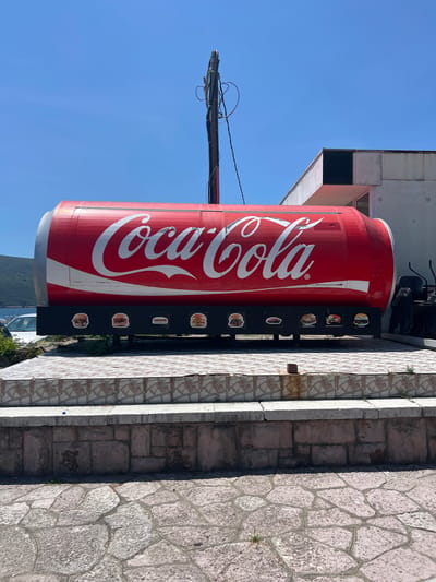 The image shows a large, cylindrical Coca-Cola can replica serving as a roadside food stand in Baošići, Montenegro. The can is predominantly red with the white Coca-Cola logo prominently displayed. Along the bottom of the can, there are several framed pictures of food items, including burgers, hot dogs, and kebabs, suggesting the types of food offered. The stand is situated on a raised platform made of stone and tiled steps. In the background, a white building with a dark square window is visible, along with stacks of chairs. Beyond that, a body of water and a lush, green hillside are present under a clear blue sky. The overall scene depicts an outdoor dining or food service establishment in a coastal area.
