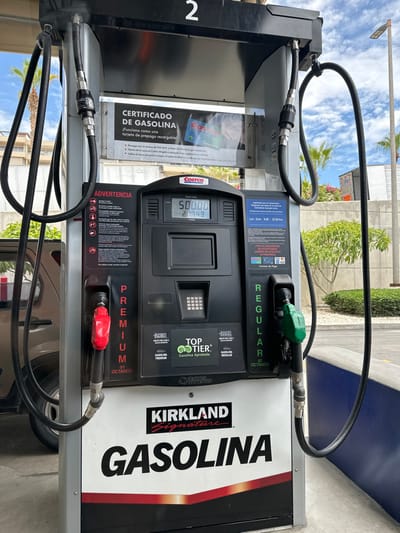 This image displays a black and silver gasoline pump, identified as pump "2," at a Costco gas station in El Tezal, Mexico. The pump features the "Kirkland Signature GASOLINA" branding at its base. Its central digital display indicates a "VENTA TOTAL" (Total Sale) of "500.00" and a dispensed volume of "21.749 LITROS." Two fuel nozzles are holstered on the pump: a red-handled nozzle on the left, labeled "PREMIUM 91 OCTANOS," and a green-handled nozzle on the right, labeled "REGULAR 87 OCTANOS." Both nozzles are connected to the pump by black hoses with silver metal fittings.

Various informational signs are present on the pump's face: a "CERTIFICADO DE GASOLINA" sign at the top detailing a reloadable prepaid card program, an "ADVERTENCIA" (Warning) section with safety pictograms on the left, and an "Importante para el consumidor" (Important for the consumer) section on the right, outlining operating hours (Monday-Sunday, 8:00-22:00 Hrs) and accepted "Formas de Pago" (Payment Methods), including Costco Visa, Costco cards, and cash. A "TOP TIER+" gasoline approval logo is also visible.

The immediate environment includes a portion of a light-colored vehicle visible to the left of the pump. The ground is paved concrete. To the right, a blue-painted concrete barrier or curb is present. The background features green foliage, including palm trees, a concrete wall, and distant buildings, all beneath a sky with scattered white clouds. No individuals are visible within the frame.