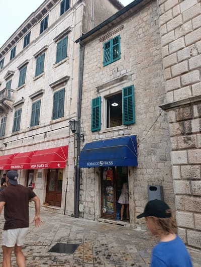 The image captures a street scene in Kotor, Montenegro, with a focus on architectural details and commercial establishments. In the foreground, a person wearing a brown t-shirt and white shorts is moving away from the camera, with their left arm extended. To the right, another person, a child, wearing a blue shirt and a black baseball cap, is walking towards the right edge of the frame.

The background features a multi-story building constructed of light-colored stone blocks. The building has numerous windows, many of which are adorned with dark green louvered shutters. On the ground floor, there are two businesses. The one on the left has a prominent red awning with "PRVA BANKA CG" printed on it. The one on the right, marked with a blue awning that reads "TOBACCO PRESS," appears to be a shop. Through the doorway of the "TOBACCO PRESS" shop, a person in a light-colored dress is visible.

A black street lamp is mounted on a pole between the two businesses. The ground is paved with cobblestones, and a metal grate is set into the pavement. The overall impression is of a historic European street, likely within the old town of Kotor, characterized by its stone architecture and narrow walkways.