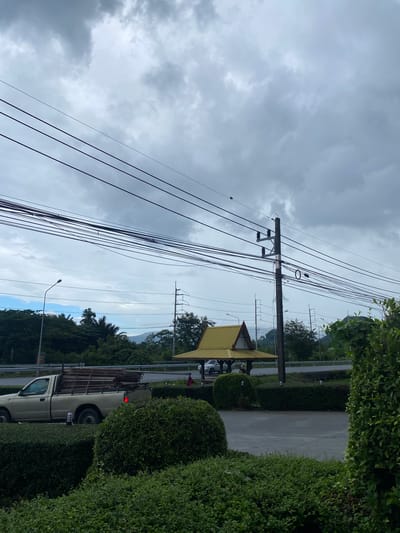 The scene is an outdoor environment in Ban Ko Pru, Thailand, showing a roadside view under a cloudy sky. The main features include power lines stretching across the sky, connected to a utility pole. Adjacent to the road is a yellow-roofed structure. A light-colored pick-up truck with construction materials in the bed is partially visible along the road. Bushes are present, lining the side of the road. The overall environment appears to be a suburban setting. No people are clearly visible. The presence of power lines and the vehicle suggest a functional environment.