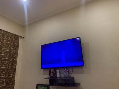 The image shows a television screen displaying a bright blue hue with a faint silhouette of a person. Below the television, a small black shelf is mounted on the wall. On the shelf, there are two trophies and a white cylindrical object, possibly an air freshener. A framed item, partially visible on the lower left, is also present. To the left of the television and shelf, a brown curtain with a wavy pattern hangs from a curtain rod. The walls are painted a light color, and a white ceiling with crown molding is visible at the top. A bright light source shines from the ceiling. The scene appears to be indoors, likely in a living room or entertainment area. There is no indication that this scene is located in Takum, Nigeria.