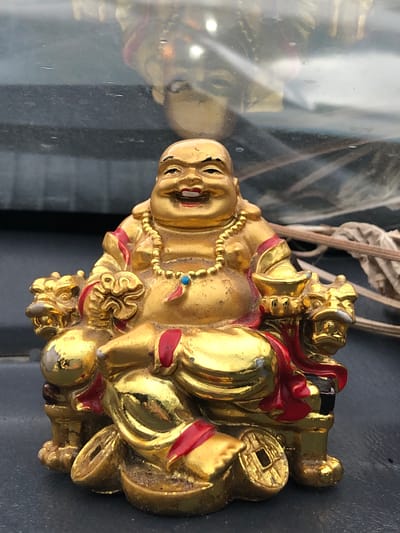 This is a close-up, slightly angled, horizontal shot of a golden, decorative figurine, likely a laughing Buddha or a similar deity. The figurine is predominantly gold-colored with accents of red and a small blue bead on a necklace. It appears to be sitting and is adorned with intricate details, including what looks like a coin with a square hole. The figurine is placed on a dark, reflective surface, possibly glass or polished stone, which shows a distorted reflection of the figurine and its surroundings. In the background, out of focus, are some dry, straw-like elements and a dark, textured surface. The lighting suggests it could be daytime, but the specific time of day, weather, or explicit location cues are not evident beyond the suggestion of an indoor setting with reflective surfaces. There is no visible text. The figurine itself seems to convey a sense of joviality and prosperity.