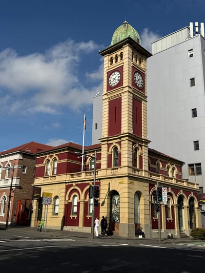 The image shows the exterior of a historic building with a prominent clock tower, identified by signs as the Redfern Post Office in Glebe, Australia. The clock tower is red and gold with a green dome. Adjacent to it is a modern, light-grey multi-story building. In the foreground, a street intersection with pedestrian crossings is visible. A traffic light shows a red signal. Several people are present: two individuals are walking away from the camera on the sidewalk, one person is sitting on the curb, and a bright green bicycle is parked on the sidewalk. The sky is blue with scattered white clouds. The building features arched windows and decorative stonework. A mural of an owl is visible on the lower portion of the clock tower.