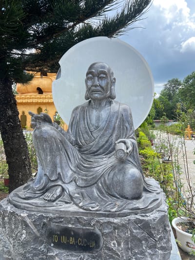 A dark grey stone statue of an elderly, bald male figure, identified by the inscription "TO UU-BA-CUC-DA" on its pedestal, is prominently displayed outdoors in the city of Đăk Mar, Vietnam. The figure is seated cross-legged, draped in robes, with a stern, wrinkled face, a beard, and elongated earlobes. His right hand extends forward with an open palm, while his left hand holds a rolled scroll on his knee. A large, smooth, light grey circular disk serves as a backdrop behind his head. To the left, a large coniferous tree partially obscures a golden-orange, multi-tiered temple structure in the background. To the right, a cultivated garden area with various green plants and small trees extends along a light-colored path. The sky above is overcast with grey clouds.