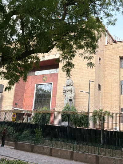 The image shows the Reserve Bank of India building in New Delhi, India. The building's facade is made of beige brick with some reddish-brown sections. A large statue of a female figure stands outside the building, to the left of a large window. A fence with barbed wire on top surrounds the property. A tree with green leaves partially obscures the top of the building. A person in a dark shirt and pants stands near a "No Stopping No Parking" sign on the left side of the frame.