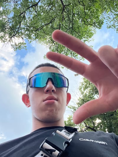 The media file is a first-person, low-angle, outdoor shot featuring a young person wearing distinctive, multi-colored, reflective sunglasses. Their face is angled upwards towards the sky, with their left hand in the foreground, fingers extended and forming a peace sign. The background consists of bright blue sky with scattered white clouds, and the lush green foliage of trees. The person is wearing a black t-shirt with a visible logo on the strap of a bag or accessory worn across their chest. The visible text on this strap reads "CALVIN KLEIN" in white, capitalized lettering.

The sun is shining brightly, casting some light through the tree branches, suggesting a daytime setting, likely late morning or afternoon given the sky and lighting conditions. The perspective and the clear sky hint at a pleasant weather day. The presence of trees and open sky could indicate a park, a garden, or an outdoor recreational area. The overall mood conveyed is casual and relaxed, possibly capturing a moment of leisure or fun. The location context provided is Petrich, Bulgaria, which aligns with the outdoor setting.
