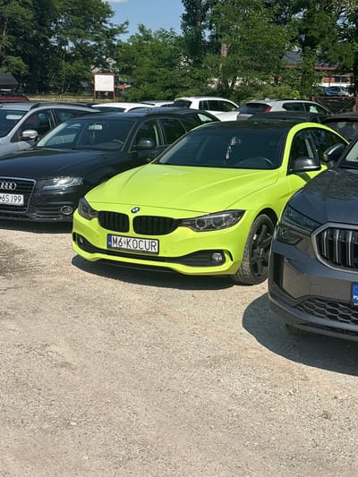 In Kobyla Góra, Poland, a bright lime green BMW coupe is parked on a gravel lot next to a black Audi sedan and a dark gray SUV. The BMW's license plate reads "M6-KOCUR" with the text "GRUPA SLIWINSKI" underneath. The Audi's license plate is partially visible, showing "6SJ99". In the background, trees and buildings are visible under a clear blue sky. The scene appears to be a daytime outdoor setting.