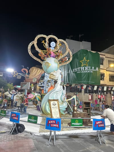 The image depicts a nighttime street scene in Alicante, Spain, likely during a festival or event. A large, elaborate artistic installation dominates the foreground, featuring a globe atop a swirling structure adorned with musical instruments like violins and trumpets. The installation is illuminated and appears to be part of a "Fallas" celebration, a traditional festival in the Valencian Community.
In the background, buildings are visible with awnings and windows. A large green banner hangs from one building displaying a yellow star and the text "ESTRELLA DE LEVANTE DE LEVANTE." Smaller banners with various designs hang below it.
Several people are present in the scene. In the mid-ground, figures are seen around the installation. In the foreground, a man in a light-colored shirt and dark shorts is leaning on a barrier, looking towards the right. Behind him, several small blue signs with red lettering "ANIS TENIS" are displayed on stands. Another sign with a green background and a yellow star says "ESTRELLA DE LEVANTE."
The ground is paved with bricks and some areas are covered in white material, possibly for display or protection. Festive lights are strung across the street in the distance, adding to the celebratory atmosphere. The overall impression is of a vibrant outdoor event in a Spanish city at night.
