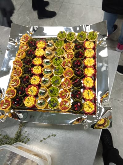 The image presents a close-up view of a large, rectangular silver serving tray, likely made of aluminum foil, laden with numerous small, artfully arranged appetizers or tartlets. The tray sits on a metallic table or countertop. Approximately 60 individual tartlets are visible, neatly arranged in rows, showcasing a diverse palette of colors and fillings.
The fillings vary significantly: some tartlets feature a reddish-brown base topped with slivered nuts (possibly pistachios or almonds), suggesting a savory spread like meat tartare or muhammara. Others contain a dark, rich filling adorned with bright red pomegranate seeds, potentially a sweet or savory compote. Bright yellow fillings, possibly made from pulses like fava beans, corn, or a pastry cream, are also prominent. Green-hued tartlets are topped with fresh herbs, such as basil or mint, or a pistachio-based spread. Another distinct variety includes white mushroom slices and a green garnish on a lighter, creamy filling. The tartlet shells themselves are varied, some appearing as simple cups, while others have decorative, flower-like edges.
In the bottom-left corner of the frame, fresh sprigs of green herbs, possibly thyme or rosemary, are scattered on the metallic surface. Adjacent to them, a clear plastic container with a white lid is partially visible, holding what appears to be finely chopped ingredients.
Several people are partially visible, indicated by their legs and feet in the background and foreground. One person in the upper left wears black shoes and pants, while another in the upper right sports blue jeans and pink/white sneakers. A third person's black shoe is visible in the bottom right. The indoor setting, with its utilitarian metallic surfaces and professionally presented food, suggests a catering event, a buffet, or a food preparation area in Riyadh, Saudi Arabia. There is no visible text in the image.