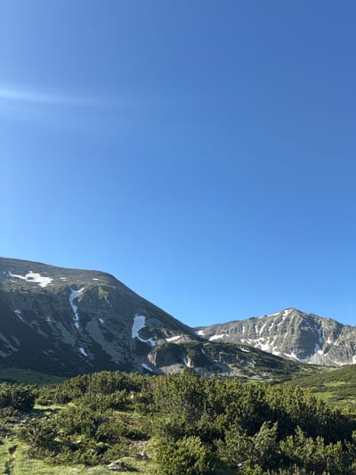 The image depicts a vast mountain landscape under a clear blue sky. In the foreground, low-lying green vegetation, likely pine bushes and grass, covers the ground. The middle ground features steep, rocky mountain slopes dotted with patches of snow. The background reveals more rugged mountain peaks, also partially covered in snow. The lighting suggests it is daytime, with a lens flare visible in the upper left corner, indicating the sun's position. The scene is natural and undeveloped, characteristic of a mountainous region. This specific image appears to be located in the Rila Mountains near Samokov, Bulgaria, a region known for its alpine scenery and hiking trails. No people or specific actions are visible in the image.