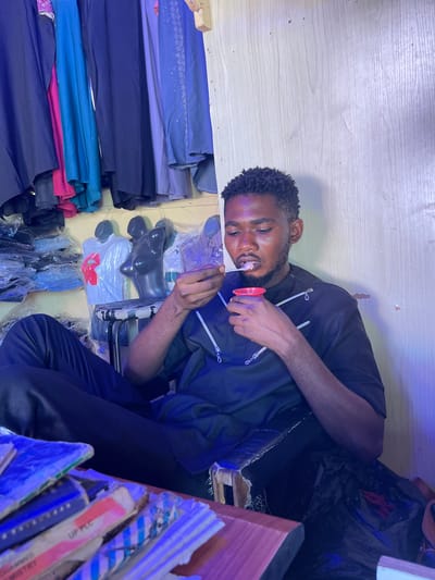 An individual is seated amidst an array of textiles and apparel in Samaru, Nigeria. The person is consuming something from a red container using a utensil. Stacks of fabrics in various colors and textures are visible to the individual's left. Apparel on mannequins, some still in packaging, is also present. To the individual's right, there are stacked items, possibly books or packaging materials. The background suggests an indoor setting, perhaps a shop or storage area.