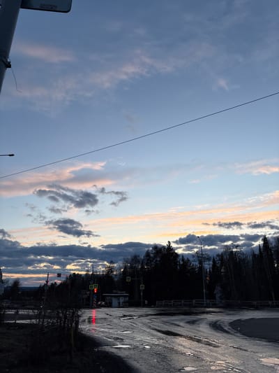 The image depicts an outdoor scene at dusk or sunset in Ust-Ilimsk, Russia, likely after recent rain given the wet, reflective road surface. The sky dominates the upper and left parts of the frame, showcasing a beautiful blend of light blue, soft pink, orange, and yellow hues, particularly where the setting sun illuminates the clouds along the horizon. Various cloud formations are present, from wispy cirrus-like clouds to more defined, layered cumulus clouds. A thin, dark cable or power line stretches diagonally across the sky.
On the right side, a dark, wet asphalt road or paved area with puddles reflects the sky's colors. The road appears worn, with visible cracks and rough patches. A dense line of silhouetted trees, possibly a forest, runs along the horizon, its dark branches stark against the colorful sky, suggesting late autumn or winter.
Amidst the trees and structures, several details are visible: a traffic light displays red signals, and a yellow diamond-shaped road sign indicates a pedestrian crossing. Behind the trees, a building or structure features a yellow sign with prominent Cyrillic text reading "ДОМ ТЕАТРА" (House of Theater). A street lamp or utility pole is also silhouetted against the sky. No people or specific activities are visible, contributing to a serene and somewhat muted atmosphere as day transitions to night.