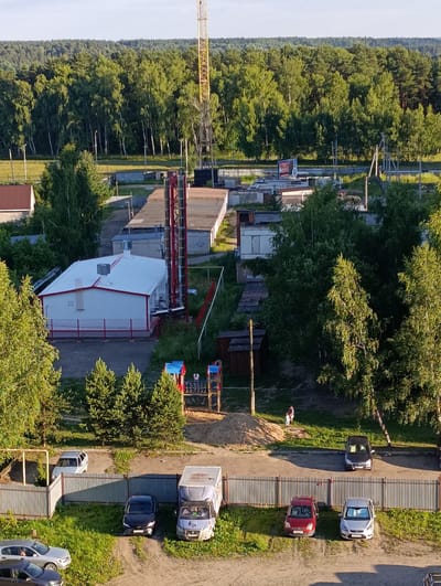 The image is an aerial view of a semi-industrial or suburban area, likely in Alexandrov, Russia, during the late afternoon or early evening, indicated by the warm, low-angle sunlight casting long shadows. The scene features a mix of buildings, vegetation, and vehicles.
Prominently in the foreground, a beige metal fence separates a dirt/sand area from a grassy patch where several cars are parked. These include a dark blue sedan, a red hatchback, and a silver hatchback. Further back, more cars are visible, including a silver hatchback and a darker sedan. A light blue van or small truck is also parked near the fence.
Behind the fence, there's a playground with a sandy base and some colorful equipment, including red and blue slides. A small child in a light-colored outfit and an adult in a dark outfit are present near the playground. A pile of sand is also visible.
Several low-rise buildings with metal or tiled roofs are scattered throughout the scene. One white building with red trim stands out, appearing to be a small industrial or utility structure. Adjacent to it, a tall red and yellow lattice tower, possibly a radio or transmission tower, rises above the buildings. A dense forest borders the area in the background, with tall green trees under a clear, light blue sky. A billboard is visible in the distance near the treeline. The overall atmosphere suggests a quiet, ordinary day in a mixed-use area.