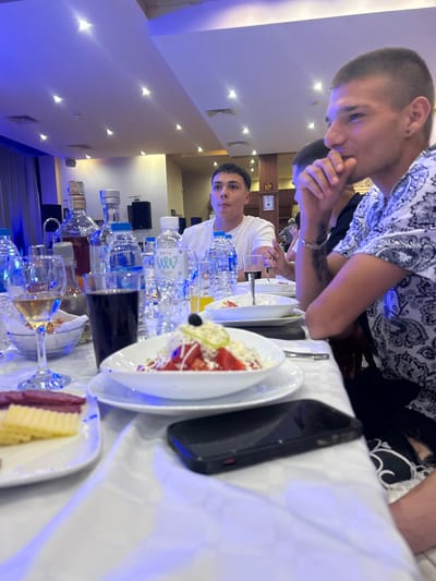 The image shows a long table laden with bottles of water, glasses, plates of food (salad and what appears to be cheese and meat), and a cell phone. Several people are seated at the table, seemingly eating or preparing to eat. The lighting suggests an indoor setting, possibly a restaurant. The presence of water bottles and the types of food indicate a meal or a gathering with refreshments. The picture was taken in a restaurant of the city Elhovo, Bulgaria.