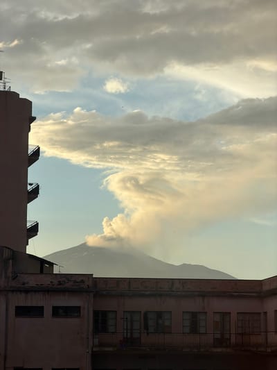 The image depicts a view of Mount Etna erupting, viewed from the city of Catania, Italy. In the foreground, the upper floors of two buildings are visible. The building on the left is tall and has multiple balconies with metal railings. The building on the right is lower and appears older, with a pink facade and many windows. The erupting volcano is in the background, with a large plume of smoke and ash rising into the sky. The sky is filled with clouds, some of which are illuminated by the sun. The overall impression is one of a dramatic natural event occurring within an urban setting.