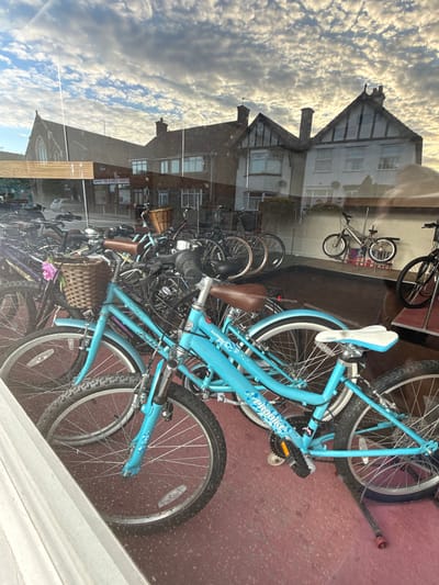 The image shows a shop window in Great Yarmouth, United Kingdom, displaying several bicycles. The bicycles are various models and colors, with one prominently being a light blue "Probike" model. The scene is layered with reflections. In the reflection, buildings are visible across the street, with a sky full of scattered clouds. The background inside the shop appears to be a red or maroon color, likely part of the shop's interior.